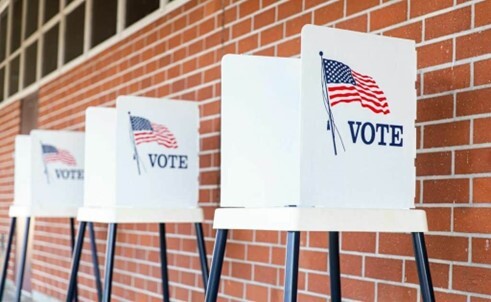 Three white stools lines up against a brick wall. There are privacy folders with the word vote and an American Flag sitting on top of the stools.